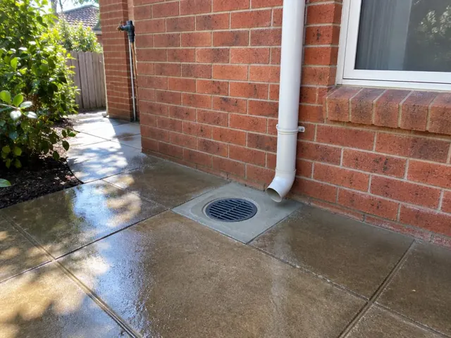 Wet paved side yard with a round outdoor drain grate beside a brick wall and downpipe in Westleigh