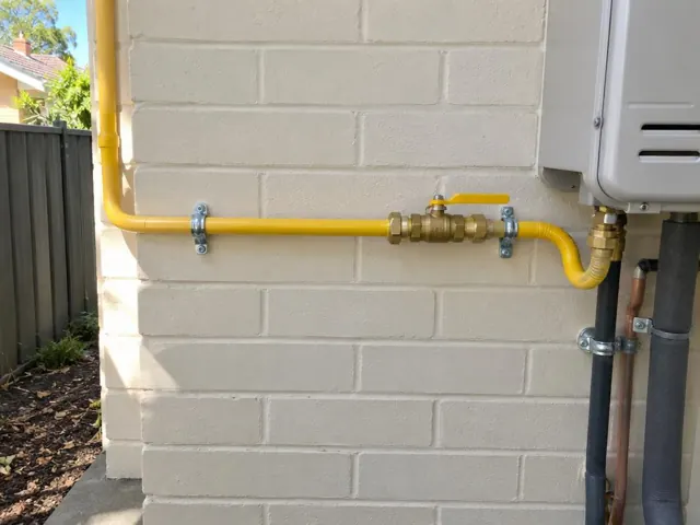 Yellow gas pipe and shutoff valve on an exterior brick wall beside a wall-mounted appliance in Thornleigh